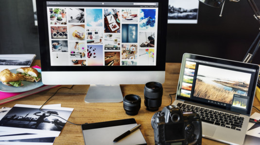 A desktop computer, a laptop, a camera, and some printed pictures on a desk.