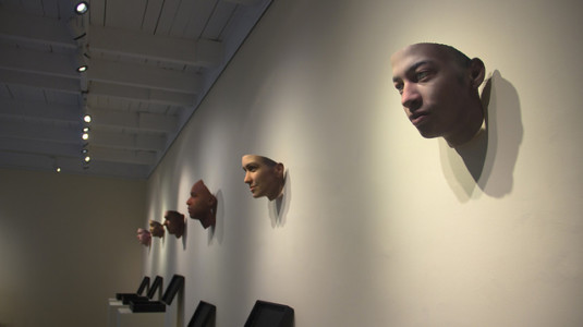 An assortment of masks hung up on the wall inside an art gallery. The masks are part of an exhibition called